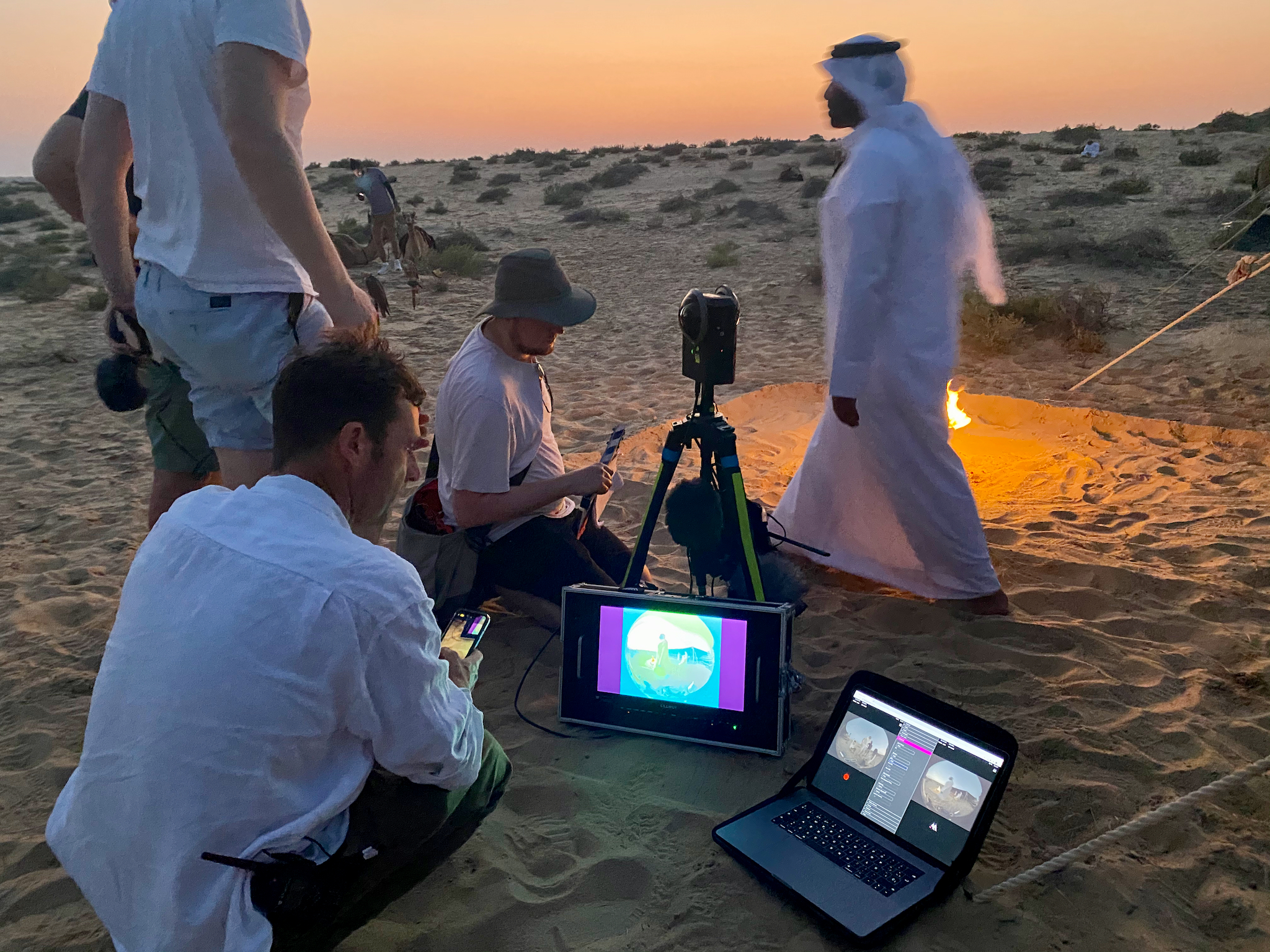 The crew at dusk in the desert, lit by firelight and a laptop screen, with the camera on a tripod.
