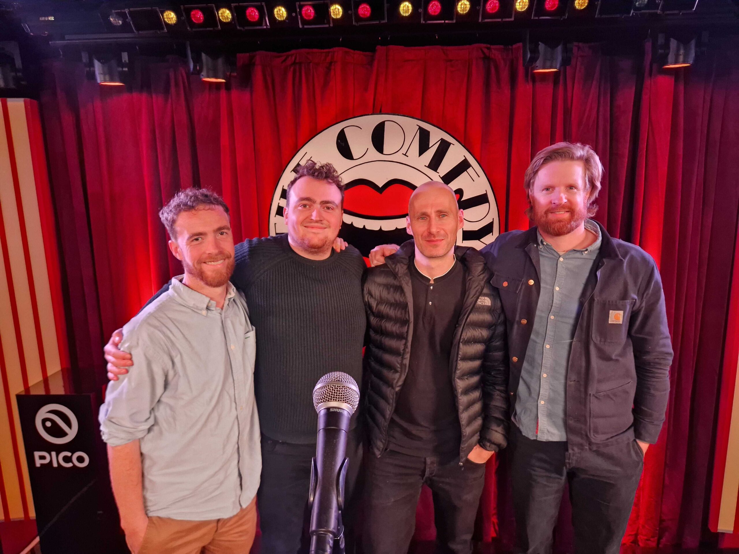 Four members of the team standing on the Comedy Store stage in front of the club logo.