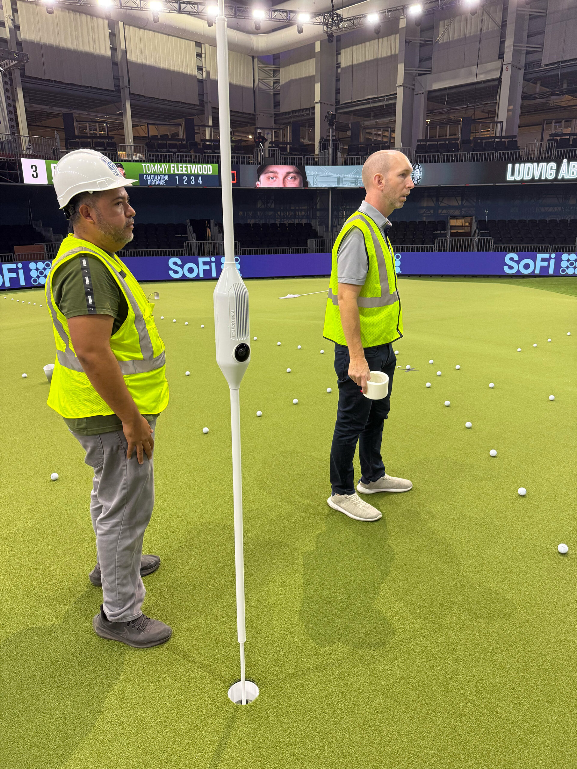 Two crew members in hi-vis jackets beside the SmartPin flagstick on an indoor green.