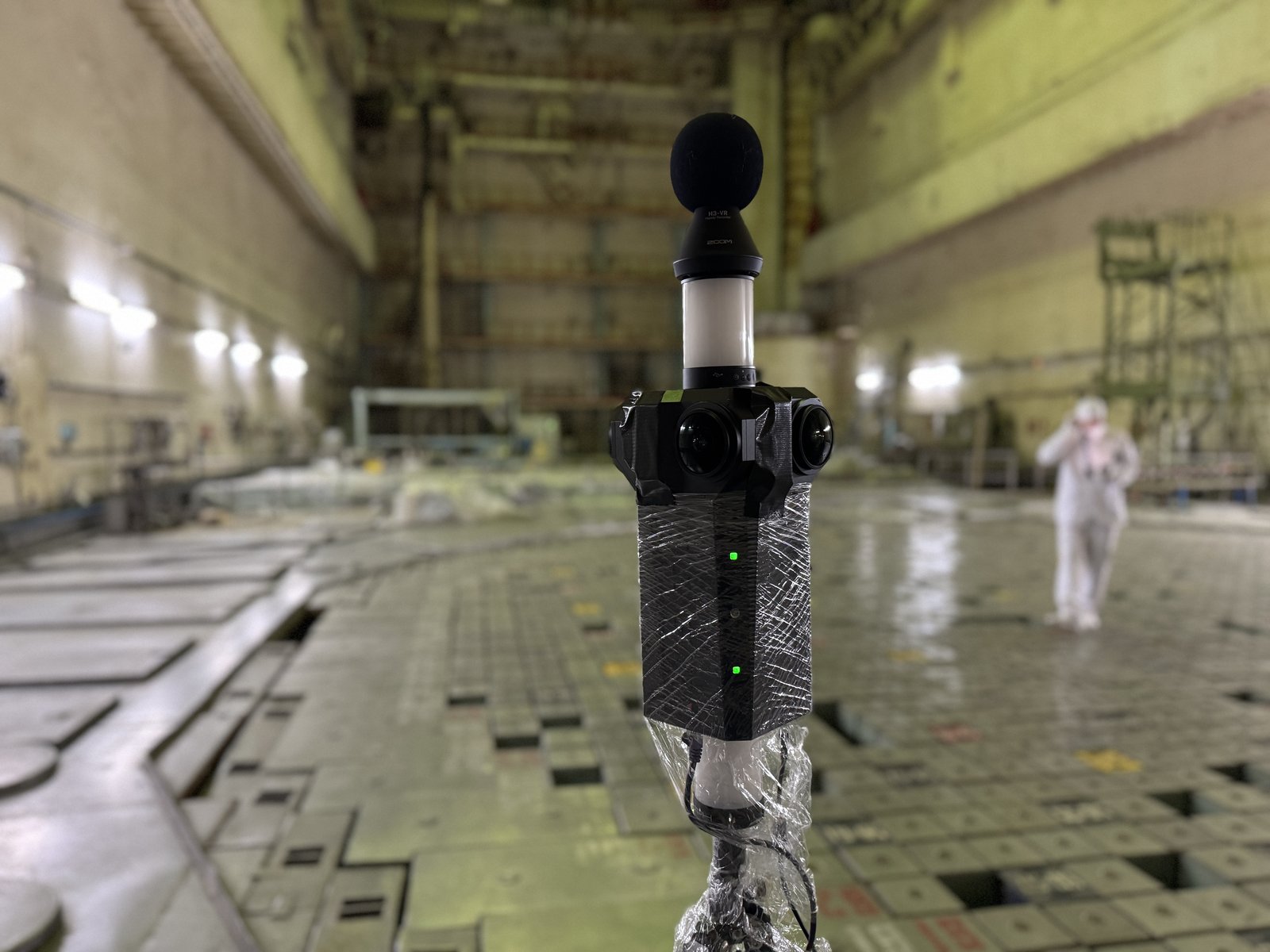 The Meta Three on a tripod inside a vast, dim industrial hall, with a figure in protective white clothing behind.