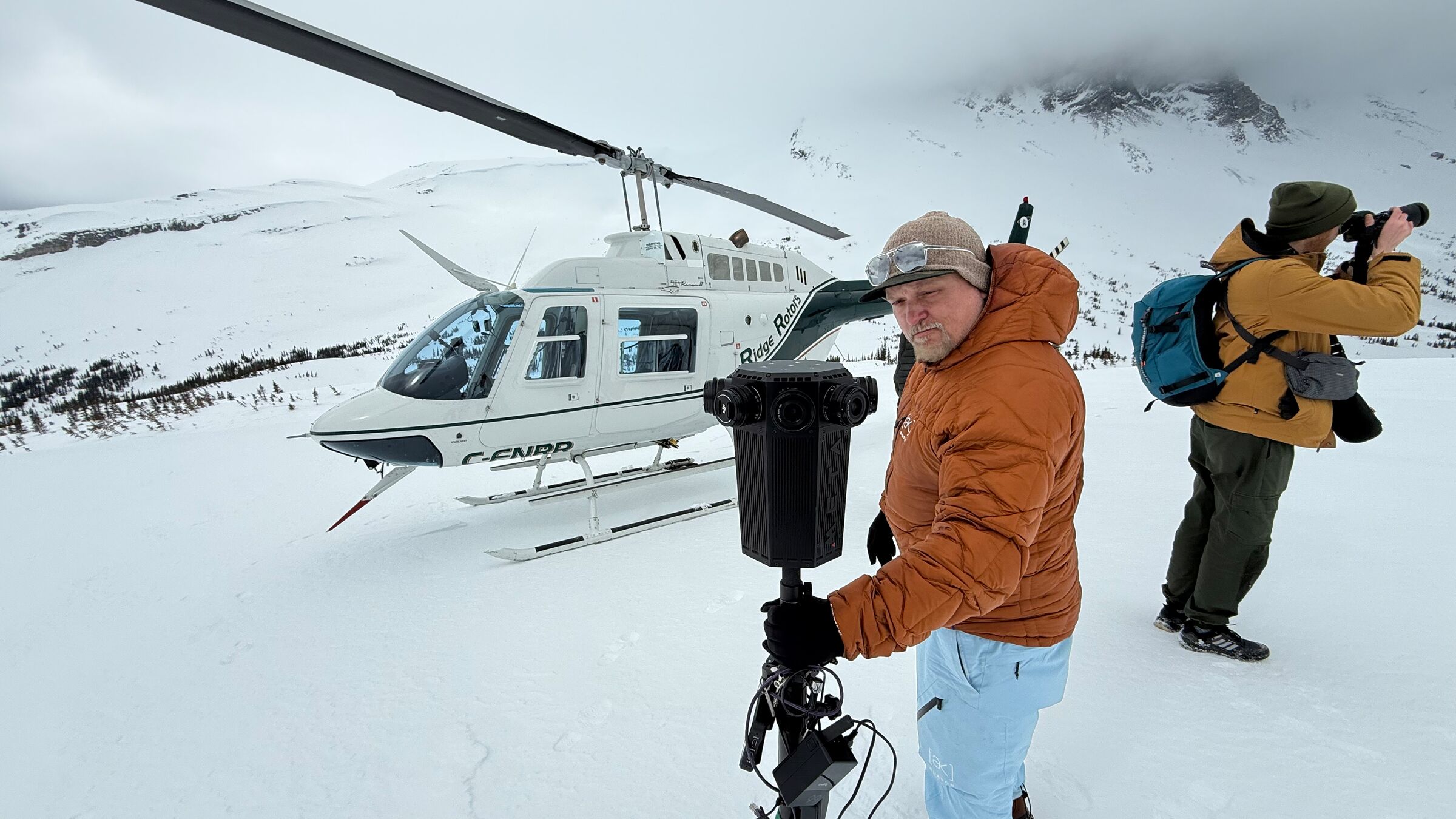 A crew member readies the Meta Four 360 camera on a snowfield beside a white helicopter.