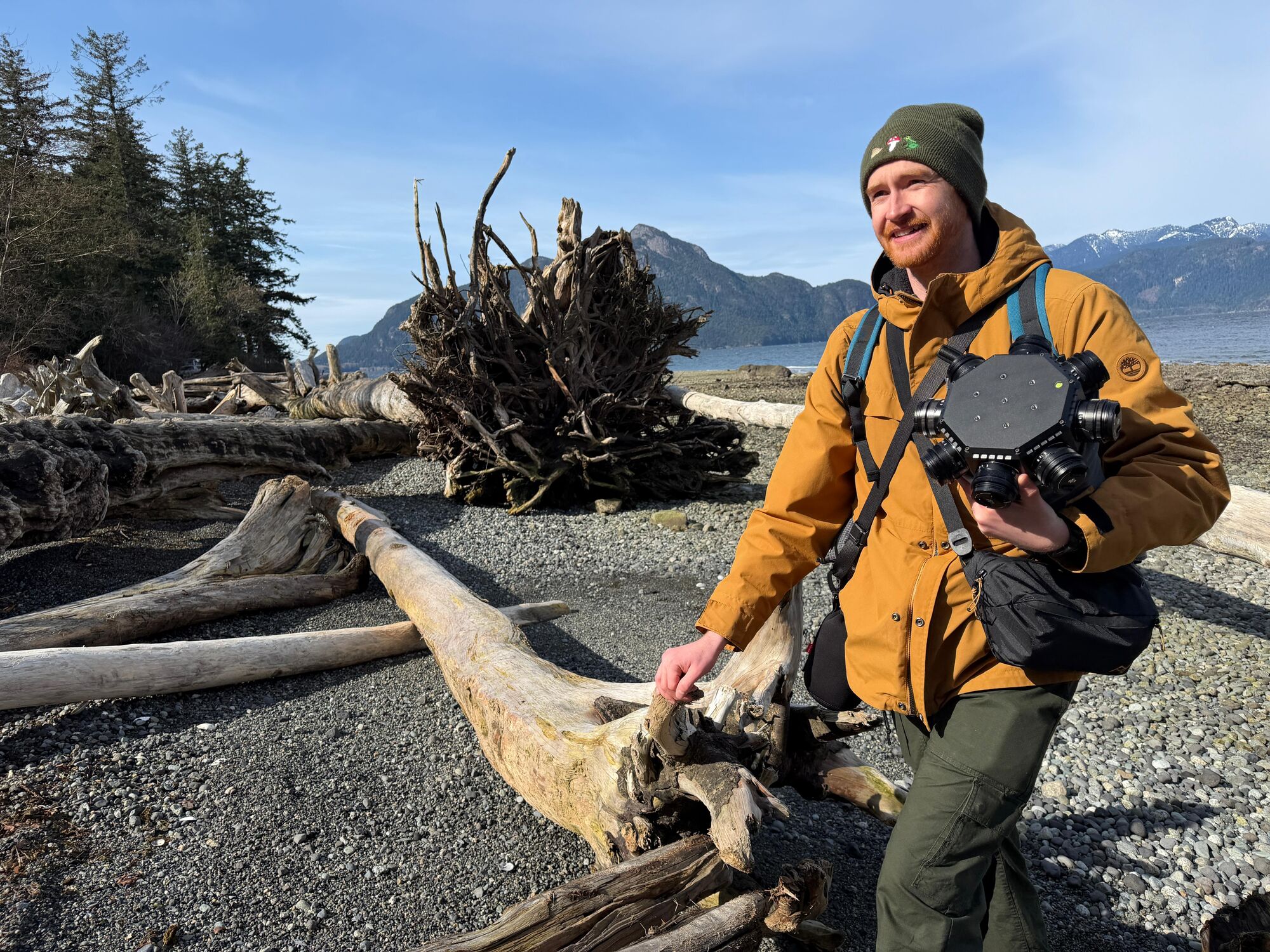 A crew member carries the camera's lens head across a beach of bleached driftwood, with mountains behind.