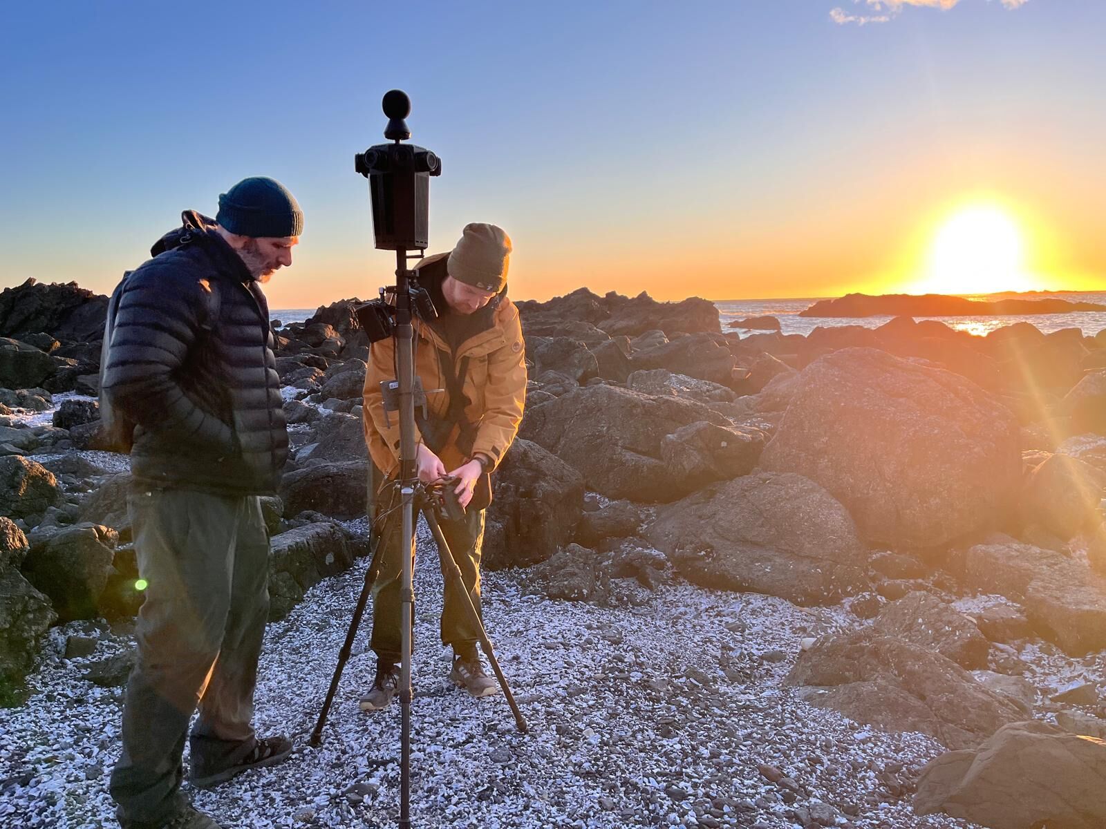 Two crew members set up the Meta Four on a tripod on a rocky, frosty shoreline at sunset.